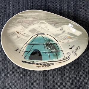 May include: Oval ceramic dish with a hand-painted mountain and igloo design. The dish features a teal igloo with a black outline, set against a backdrop of gray mountains and a light gray sky. The artist's signature, "SASCHA B", is visible.