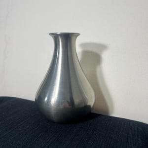 May include: A brushed metal vase with a teardrop shape and a narrow neck. The vase has a smooth, reflective surface and sits on a dark blue fabric. The vase is a decorative item.