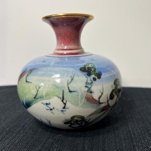 May include: A small, round ceramic vase with a landscape design. The vase has a red neck with a gold rim and a blue and white body with painted trees and hills. The vase is approximately 5 inches tall.