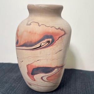 May include: A ceramic vase with a matte, light beige base color featuring swirling, marbled patterns in shades of terracotta orange, deep black, and dark brown. The vessel has a rounded body, a narrow neck, and a simple, smooth rim opening.