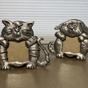 May include: Two pewter-colored picture frames, one shaped like a cat and the other like a dog. The cat frame has a curved tail and the dog frame has floppy ears. Both frames have a rectangular opening for a photo.