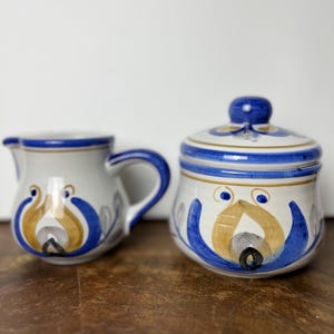 May include: A ceramic creamer and a lidded sugar bowl featuring a hand-painted folk art pattern in blue, gold, and black on a cream-colored background. The creamer has a curved handle and spout, while the sugar bowl includes a matching lid.