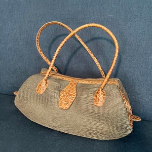 May include: A structured handbag featuring a dark gray woven mesh body with a curved silhouette. The bag includes tan, crocodile-embossed leather trim, two matching thin shoulder straps, and a central leather tab closure with metal hardware.