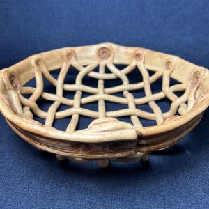 May include: A round, beige ceramic bowl featuring an openwork lattice design. The structure is composed of interconnected clay strips forming a grid pattern, with a solid, slightly raised rim that has small circular indentations along the edge.