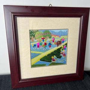 May include: A framed embroidery artwork depicting a rural scene with figures working in green fields and blue water channels. The scene features people in colorful clothing, a brown animal, and trees, all enclosed in a dark wood frame with a cream mat.