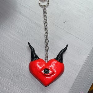 May include: A red heart-shaped charm with two black horns and a single eye in the center. The charm is attached to a silver-colored metal chain with a lobster claw clasp at the top. The surface of the heart is smooth and glossy in appearance.
