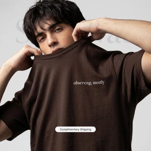 以下が含まれることがあります： 白文字で「observing, mostly」と書かれた茶色のTシャツ。Tシャツは無地で、クルーネックです。画像には「Complimentary Shipping」というテキストも含まれています。