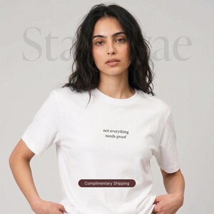 以下が含まれることがあります： ミニマリストフォントで「not everything needs proof」のテキストが入った白いクルーネックTシャツ。柔らかい素材でできており、半袖です。モデルがシャツを着ています。