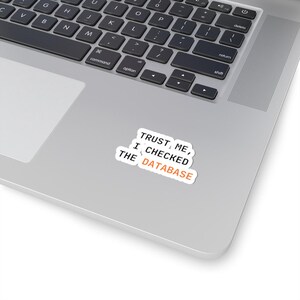 Op de afbeelding: Een witte sticker met de tekst "TRUST ME, I CHECKED THE DATABASE" in zwart en oranje. De sticker zit op een laptop, met het toetsenbord zichtbaar.