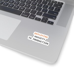 May include: A white sticker with the text "DEPLOYING ANXIETY TO PRODUCTION" in black and orange. The sticker is on a silver laptop, near the keyboard.