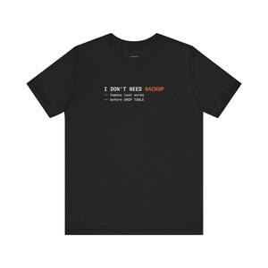 May include: Black t-shirt with white and orange text. The text reads "I DON'T NEED BACKUP" with the phrases "-- famous last words" and "-- before DROP TABLE" below it. The shirt is a short-sleeved crew neck.