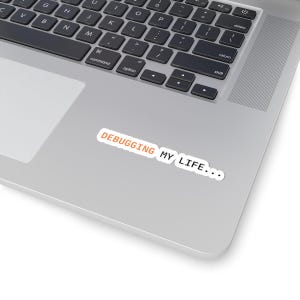 May include: A white sticker with the text "DEBUGGING MY LIFE..." in orange and black lettering is placed on a silver laptop. The laptop keyboard is visible in the background, with black keys and a silver frame.