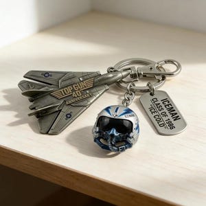 Könnte beinhalten: Ein Top Gun 40th Anniversary Schlüsselanhänger-Set. Das Set enthält ein silberfarbenes Kampfflugzeug, einen Miniatur-Pilotenhelm und einen Anhänger mit der Aufschrift "ICEMAN CLASS OF 1986 'ICE COLD'".
