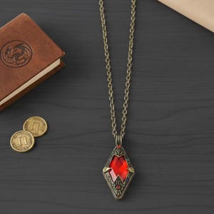 Könnte beinhalten: Eine Halskette im Vintage-Stil mit einem rautenförmigen Anhänger mit einem großen, facettierten roten Edelstein. Der Anhänger ist von kunstvollen Metallarbeiten umrahmt und hängt an einer langen, antikgoldfarbenen Kette. Ein ledergebundenes Buch und Goldmünzen sind ebenfalls sichtbar.