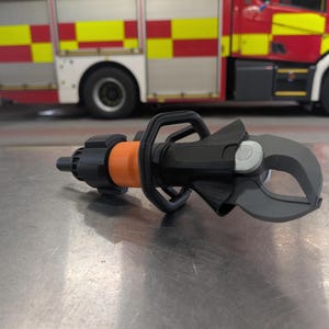 May include: A black and orange hydraulic rescue tool with grey blades, designed for emergency response. The tool is on a metal surface, with a red and yellow fire engine in the background.