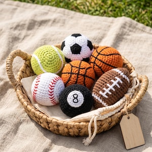May include: A woven basket filled with handmade crochet sports balls. The collection includes a tennis ball, soccer ball, basketball, baseball, eight ball, and football. The balls are in various colors, including green, white, orange, black, and brown.