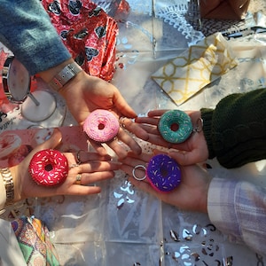 Peut inclure: Quatre mains tenant des porte-clés en forme de donut colorés. Les porte-clés sont dans des tons de rouge, rose, vert et violet, avec des détails de couture blancs. Sur une surface en plastique transparent.