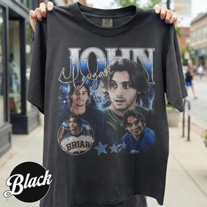 May include: Black t-shirt featuring a graphic of a man with the name "John Logan" in a vintage style. The design includes multiple images of the man, stars, and the word "Briar".