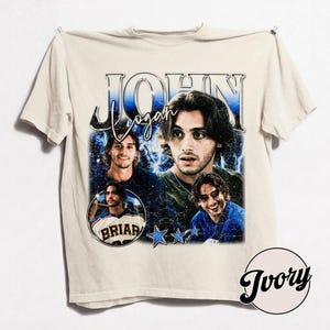 May include: Ivory-colored t-shirt featuring a graphic design with the name "John Logan" in a vintage style. The design includes multiple images of a person, blue stars, and the word "Briar" in a circle.