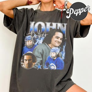 May include: A charcoal gray short-sleeve t-shirt featuring a graphic print of John Tucker. The design includes three images of the character, the text "JOHN" in large block letters, "tucker" in script, and three blue star icons with lightning.