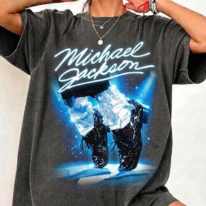 May include: Dark gray t-shirt featuring the name "Michael Jackson" in white script. The graphic shows a blue spotlight illuminating black shoes with white socks, with a starry background. The shirt has short sleeves.