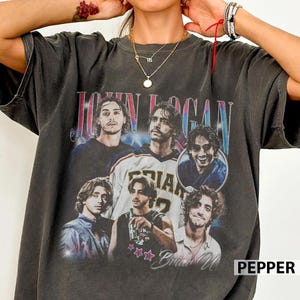 Peut inclure: T-shirt gris foncé avec un collage d'images d'un homme aux cheveux longs et ondulés. Le t-shirt porte l'inscription "JOHN LOGAN" dans une police rétro, avec le nom "Brian U" en dessous des images. Le mot "PEPPER" est dans le coin inférieur droit.