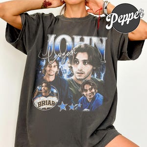 May include: Charcoal grey t-shirt with a graphic design featuring a man's image and the name "John Logan" in blue and white. The design includes multiple images of the man, the word "Briar", and blue star graphics.
