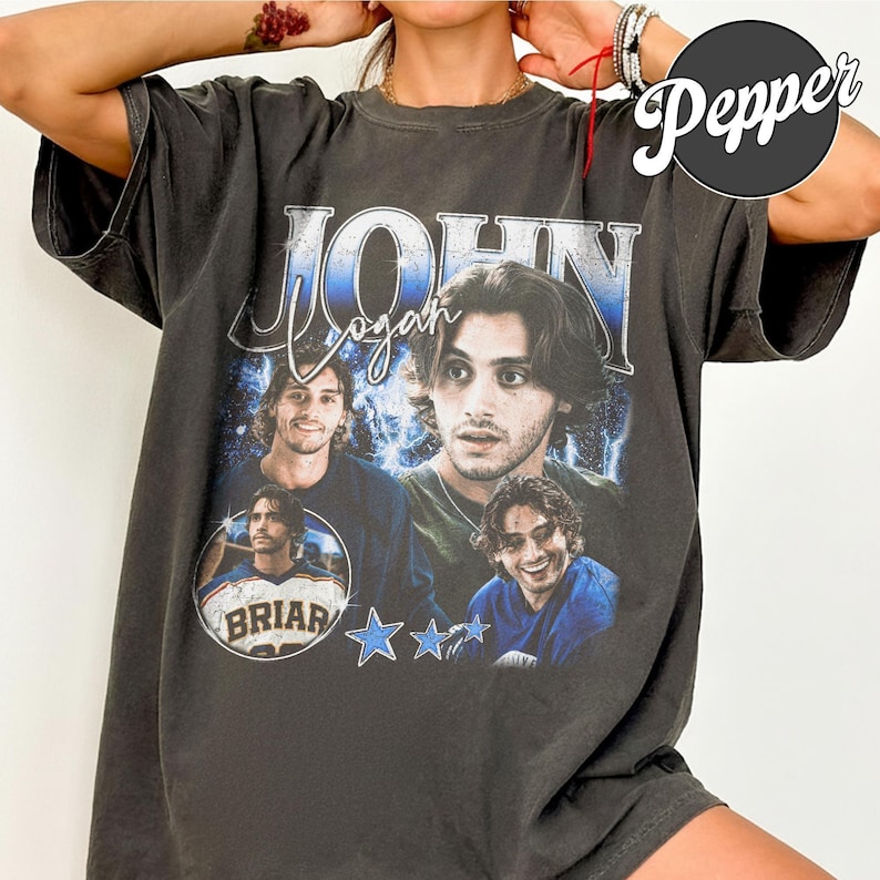 May include: Dark gray t-shirt featuring a graphic of a man with the name "John Logan" in a blue and white design. The shirt includes multiple images of the man and the word "Briar" on a circular emblem, along with blue star graphics.