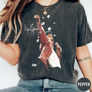 May include: Dark gray t-shirt featuring a graphic of a singer in a red jacket, holding a microphone and raising an arm. The singer's signature is above the image. The shirt is a vintage style.