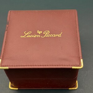 May include: A square, burgundy-colored jewelry box with gold-tone metal corner accents. The top lid features the gold-colored text Lucien Piccard with an LP logo above the name. The box has a smooth, leather-like finish and a hinged lid design.