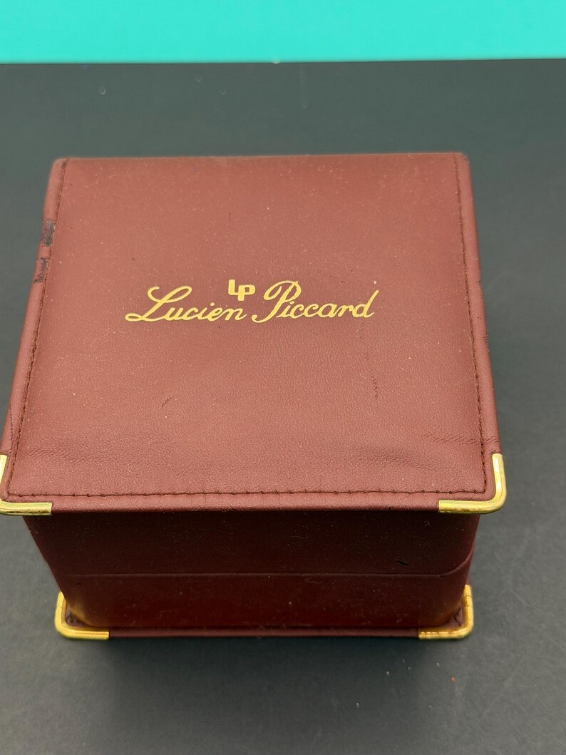 May include: A square, burgundy-colored jewelry box with gold-tone metal corner accents. The top lid features the gold-colored text Lucien Piccard with an LP logo above the name. The box has a smooth, leather-like finish and a hinged lid design.