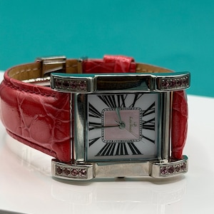 May include: A rectangular wristwatch with a silver-tone metal case and a bright red textured leather strap. The watch face features a white background with black Roman numerals, a pink center square, and small pink stones set into the lugs.