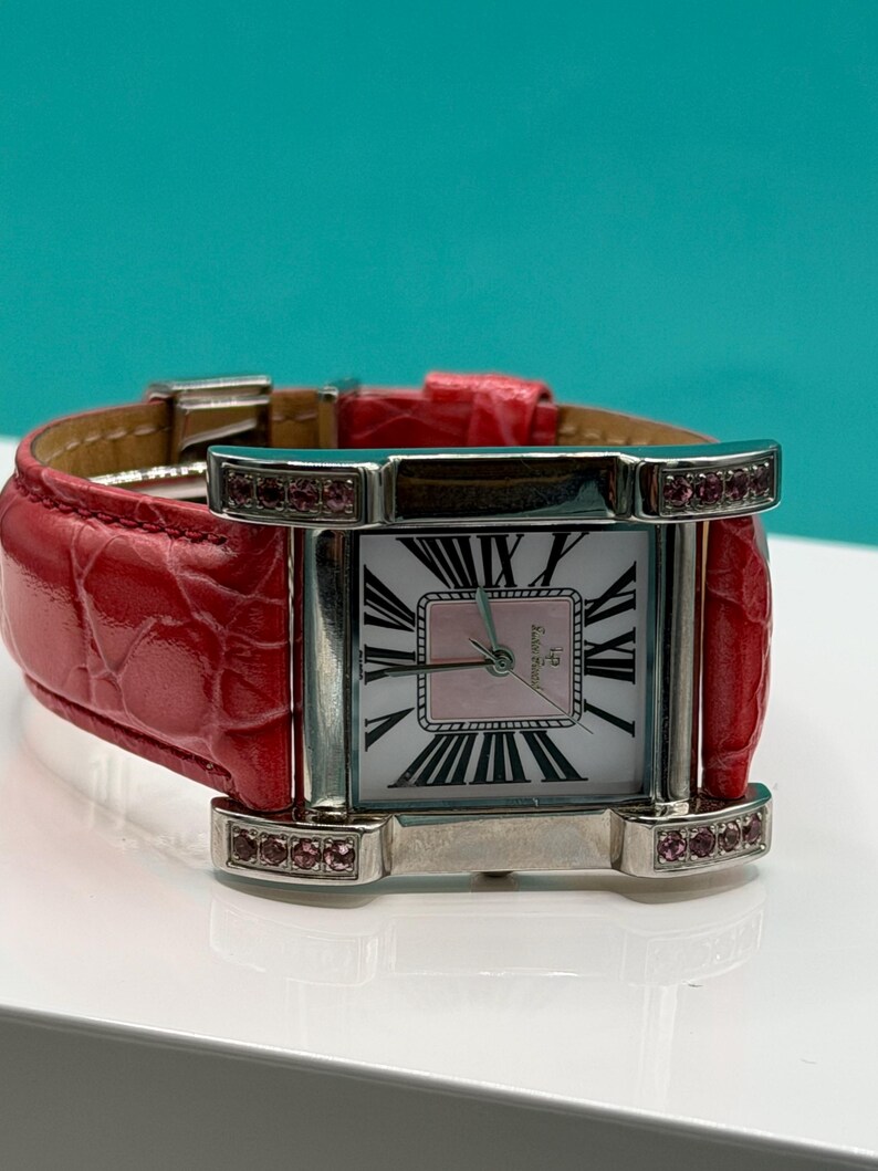 May include: A rectangular wristwatch with a silver-tone metal case and a bright red textured leather strap. The watch face features a white background with black Roman numerals, a pink center square, and small pink stones set into the lugs.
