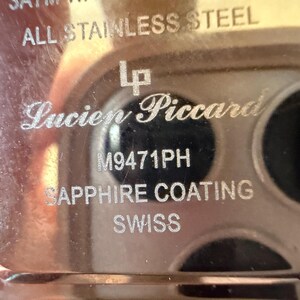 May include: The metallic back casing of a Lucien Piccard watch featuring engraved text that reads ALL STAINLESS STEEL, the LP logo, M9471PH, SAPPHIRE COATING, and SWISS. The surface is reflective and silver-toned with visible etched lettering.