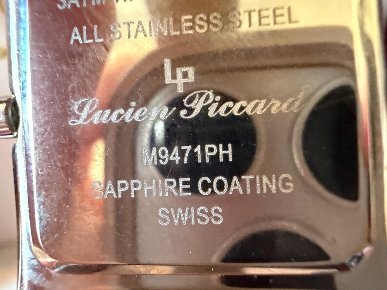May include: The metallic back casing of a Lucien Piccard watch featuring engraved text that reads ALL STAINLESS STEEL, the LP logo, M9471PH, SAPPHIRE COATING, and SWISS. The surface is reflective and silver-toned with visible etched lettering.