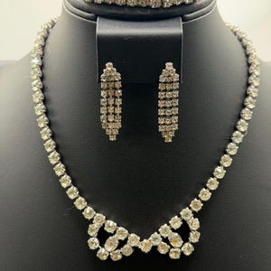 May include: A sparkling jewelry set featuring a necklace, bracelet, and earrings. The necklace and bracelet are composed of multiple rows of clear, round rhinestones. The earrings are rectangular and also feature clear rhinestones. The jewelry is displayed on a black stand.