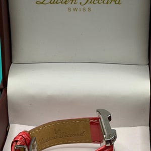 May include: A red leather watch strap with a textured finish and a silver-tone metal clasp, displayed inside a presentation box. The interior of the strap and the box lid feature the Lucien Piccard brand name and logo in gold lettering.