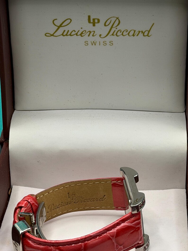 May include: A red leather watch strap with a textured finish and a silver-tone metal clasp, displayed inside a presentation box. The interior of the strap and the box lid feature the Lucien Piccard brand name and logo in gold lettering.