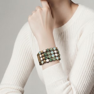 May include: A multi-row bracelet with a bronze-colored metal frame. The bracelet is adorned with rows of round, light green, white, and light blue stones. The bracelet is worn on a wrist.