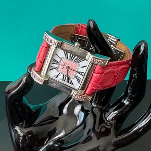 May include: A square-faced wristwatch with a silver-tone metal case, featuring a white dial with black Roman numerals and a pink center square. The watch has a bright pink textured leather strap and small pink crystals embedded in the case lugs.