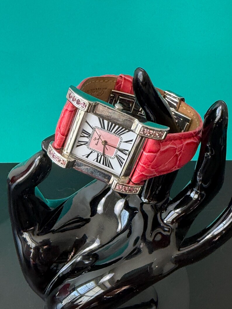May include: A square-faced wristwatch with a silver-tone metal case, featuring a white dial with black Roman numerals and a pink center square. The watch has a bright pink textured leather strap and small pink crystals embedded in the case lugs.
