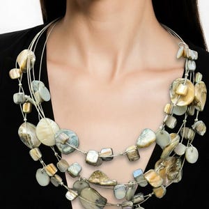 May include: A multi-strand necklace featuring a variety of natural, irregularly shaped beads in shades of beige, gray, and iridescent white. The beads are strung on thin, silver-colored wire, creating a layered effect. The necklace is a statement piece.