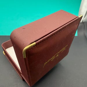 May include: A rectangular, burgundy-colored jewelry box with a hinged lid. The exterior features gold-toned metal corner accents and the gold-printed text Lucien Piccard on the top. The interior is lined with a white material.