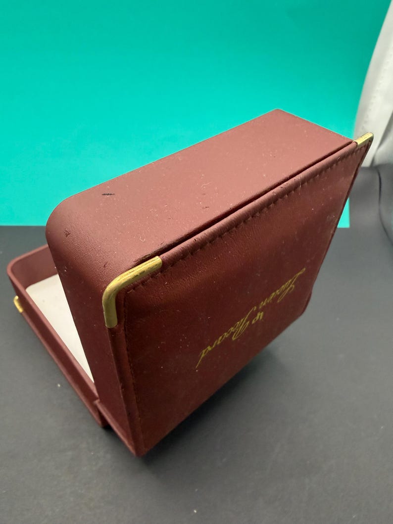 May include: A rectangular, burgundy-colored jewelry box with a hinged lid. The exterior features gold-toned metal corner accents and the gold-printed text Lucien Piccard on the top. The interior is lined with a white material.