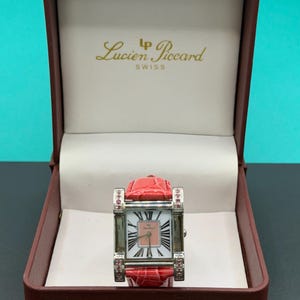 May include: A Lucien Piccard Swiss wristwatch with a rectangular silver-tone case, a pink dial featuring black Roman numerals, and a red textured leather strap. The watch is presented in a brown hinged box with the brand name printed in gold.