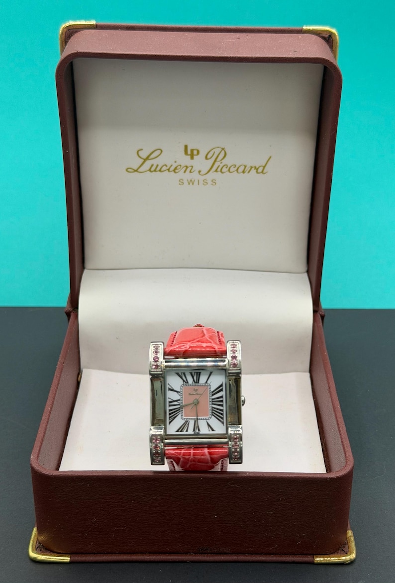 May include: A Lucien Piccard Swiss wristwatch with a rectangular silver-tone case, a pink dial featuring black Roman numerals, and a red textured leather strap. The watch is presented in a brown hinged box with the brand name printed in gold.