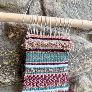 May include: A woven wall hanging with a bamboo rod and white string. The textile features horizontal stripes in shades of pink, teal, burgundy, and white. The bottom edge has a fringe with decorative wooden beads.