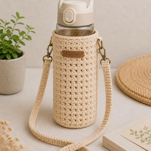 May include: A beige crocheted water bottle holder with a matching strap and a clear glass bottle. The holder has a small brown leather tag that reads "Handmade". The bottle has a beige lid with a handle. The strap is adjustable and attaches to the holder with bronze-colored clasps.