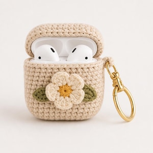May include: Beige crochet AirPods case with a white flower and green leaves applique. The case has a gold-colored clasp and holds white AirPods. The case is designed to protect and carry the wireless earbuds.