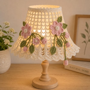 May include: A small, decorative table lamp with a cream-colored, crocheted lampshade. The shade is adorned with pink crocheted flowers and green leaves. The lamp has a wooden base and stem, and is sitting on a wooden surface.
